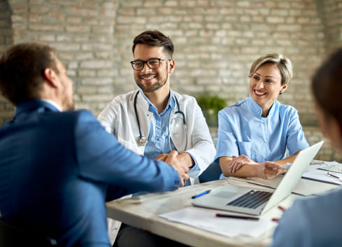 Médico y enfermera sonríen mientras estrechan manos con un hombre en una mesa. Se observa un ordenador portátil y documentos en un entorno de oficina con paredes de ladrillo.