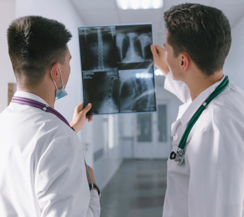 Dos médicos en un pasillo de hospital observan una radiografía en una hoja. Ambos llevan batas blancas y estetoscopios, el ambiente es clínico y luminoso.