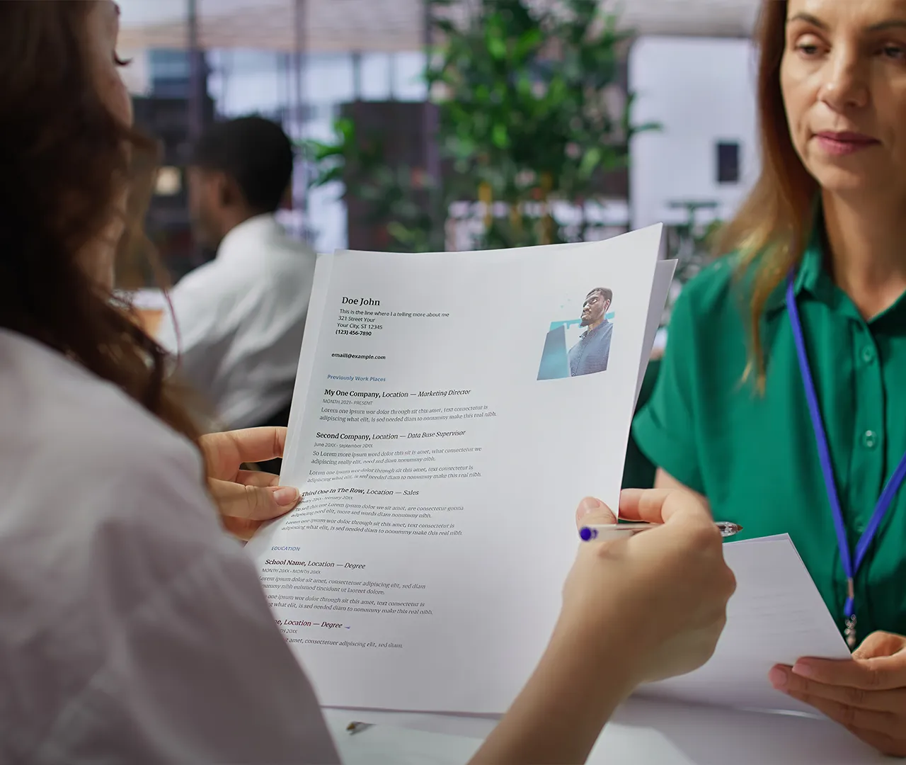 Una persona sostiene un currículum mientras otra lo revisa. Ambas están en un entorno de oficina con plantas. Predominan colores neutros y la vestimenta es formal.