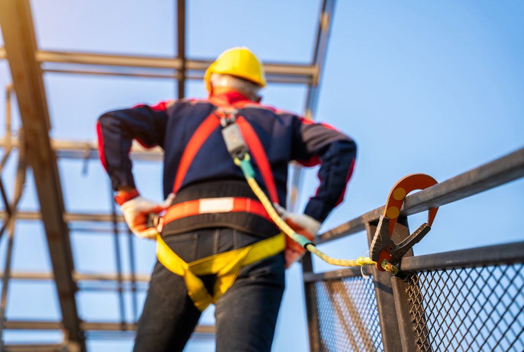 Trabajador en altura con casco amarillo y arnés de seguridad, sujetando una línea de vida en un entorno de construcción, con estructura metálica y cielo azul de fondo.