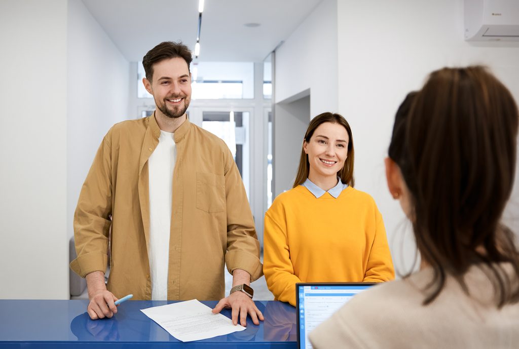 Pareja de personas sonrientes en un mostrador, una mujer con suéter amarillo y una blusa azul, un hombre con chaqueta beige. Fondo minimalista y moderno, con pantalla visible.