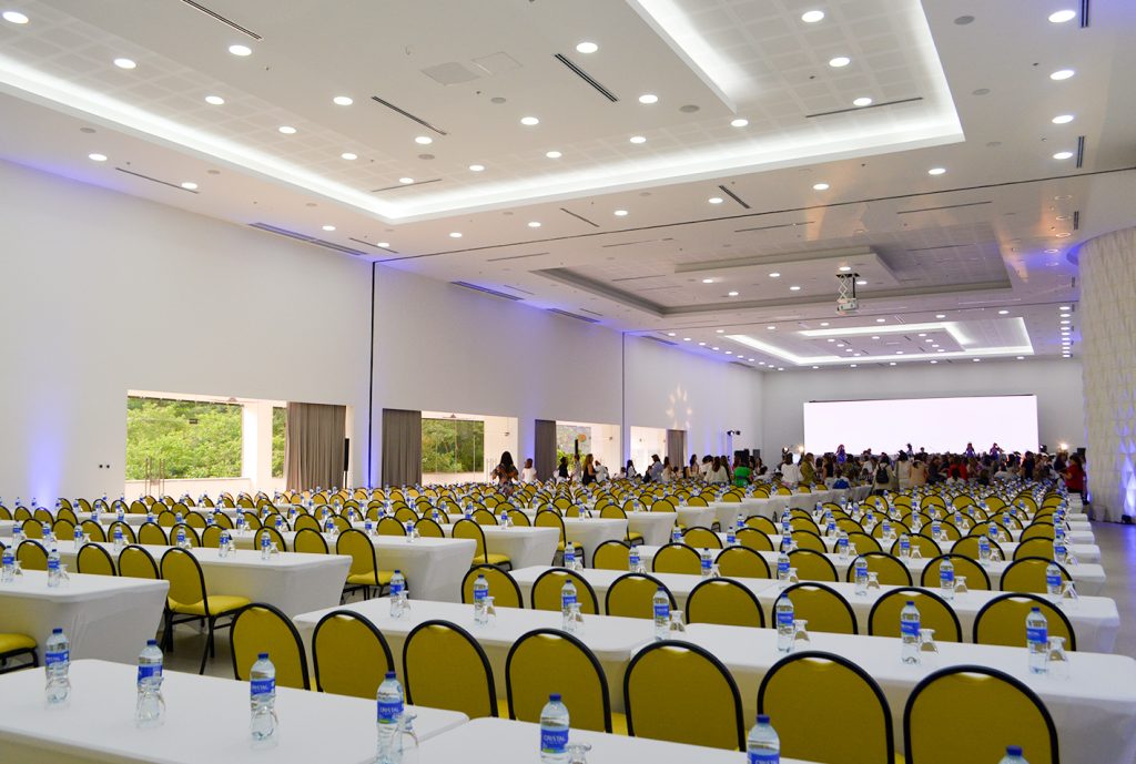 Amplio salón de eventos con mesas blancas y sillas amarillas, decorado con luces. Al fondo, un grupo de personas se prepara para el evento, con un proyector visible.