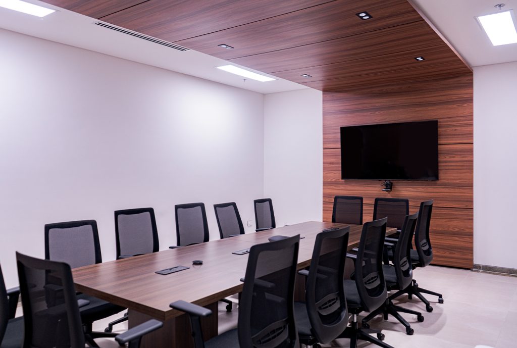 Sala de reuniones con mesa larga de madera y sillas negras. Pared de madera y pantalla plana en el fondo. Iluminación suave y ambiente moderno.