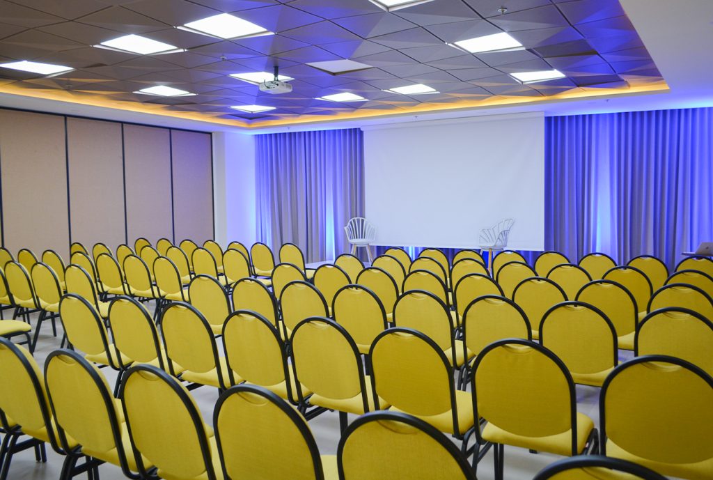 Salón de eventos con filas de sillas amarillas, un proyector en el techo, una pantalla blanca al frente y cortinas grises iluminadas con luz azul.