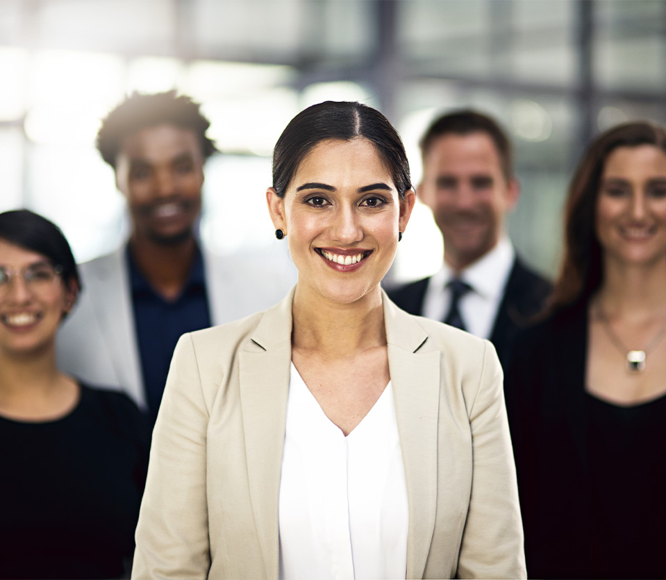 Grupo de cinco personas en un entorno profesional, con una mujer al frente sonriendo. Predominan colores claros y oscuros en la vestimenta, con un fondo desenfocado.