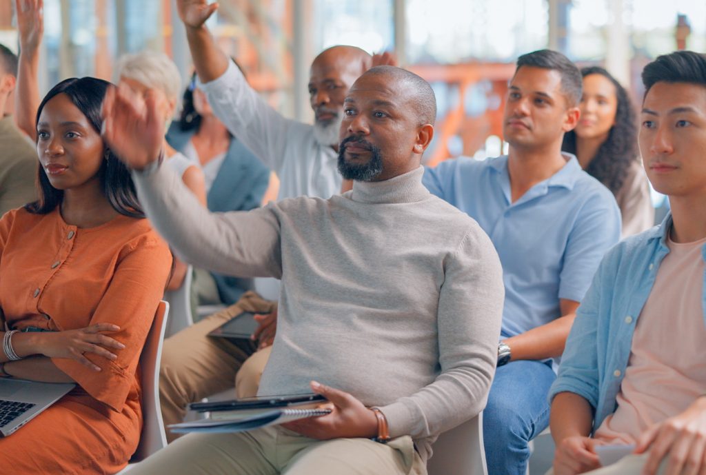 Grupo diverso de personas en un evento, algunas levantando la mano. Se observan colores neutros en la vestimenta y un entorno luminoso con ventanas grandes.