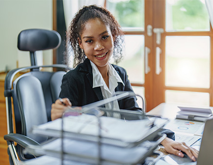 Mujer sentada en una oficina, sonriendo y sosteniendo un organizador de documentos. El entorno incluye una silla de oficina, papeles y ventanas con luz natural.
