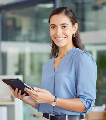 Mujer sonriente con blusa azul sostiene una tablet en un entorno de oficina moderna, con paredes de vidrio y mobiliario contemporáneo. El ambiente es luminoso y profesional.
