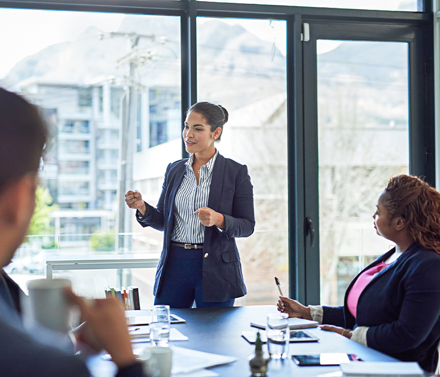 Mujer de pie en una sala de reuniones, hablando con confianza. Tres personas sentadas alrededor de una mesa, con documentos y tazas. Ventanas grandes muestran un paisaje urbano.
