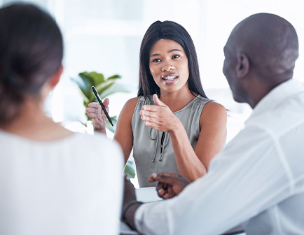 Mujer habla con gestos a dos personas en una reunión, rodeada de luz natural y plantas.