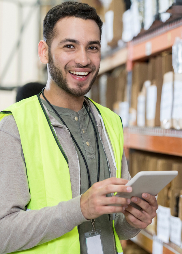Hombre sonriente con chaleco reflectante y camiseta gris, sostiene una tablet en un almacén con estanterías llenas de cajas y etiquetas visibles.