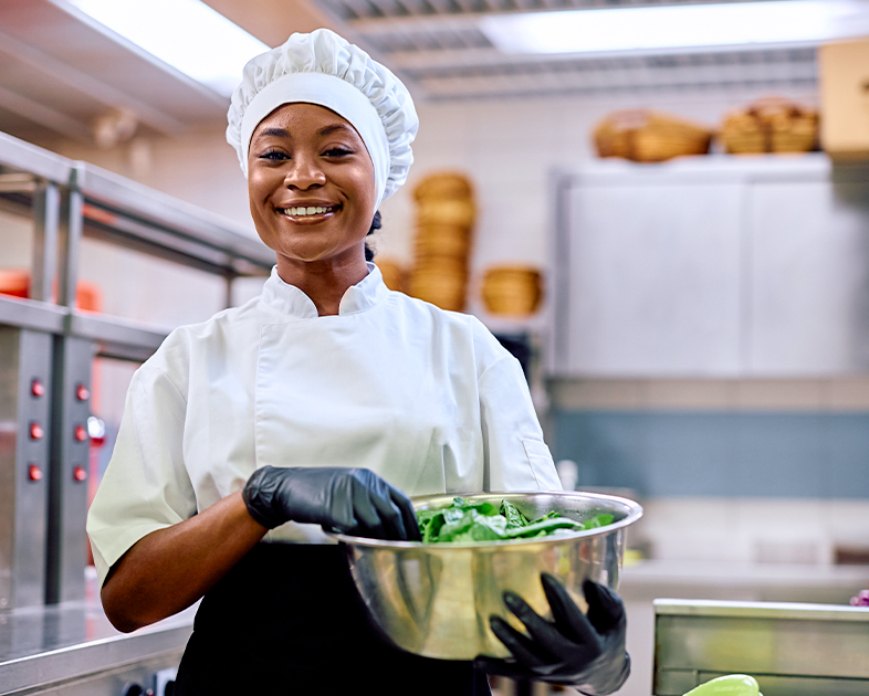 Chef sonriente sostiene un bol con verduras en una cocina. Lleva uniforme blanco y guantes negros, con un entorno de acero inoxidable y cestas en el fondo.