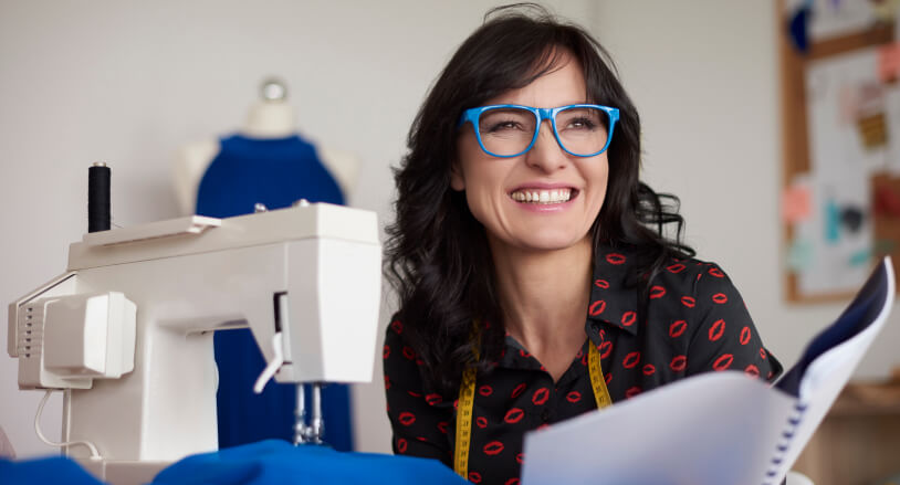 Mujer sonriente con gafas azules, sentada junto a una máquina de coser, sosteniendo un cuaderno, con una prenda de tela azul y un maniquí al fondo.