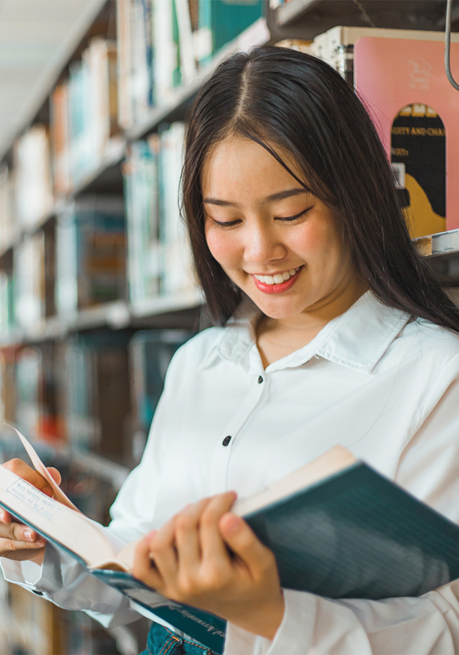 Mujer sonriendo mientras sostiene un libro en una biblioteca, rodeada de estantes con libros de diversos colores y tamaños. La luz es suave y el ambiente es acogedor.
