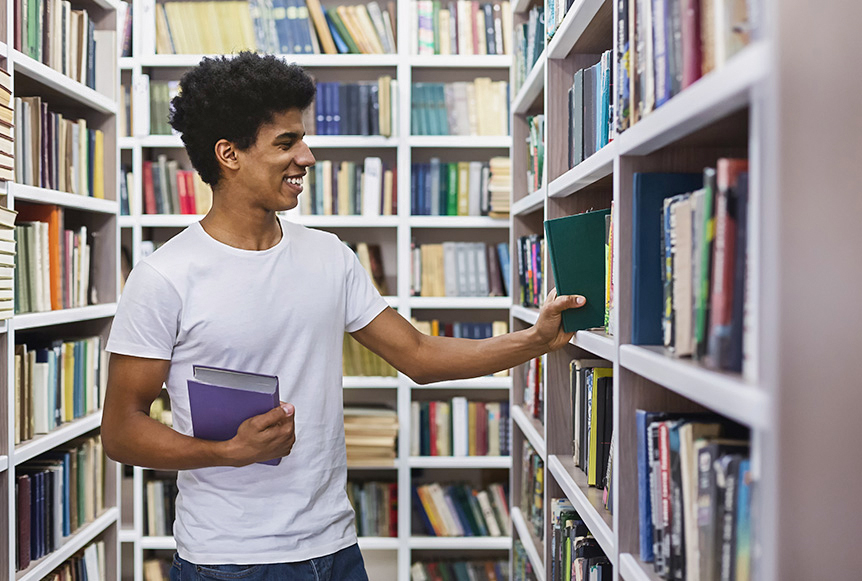 Joven con camiseta blanca sostiene libros en un pasillo de biblioteca, rodeado de estanterías llenas de libros de diversos colores y tamaños. Sonriente, examina un libro en la estantería.
