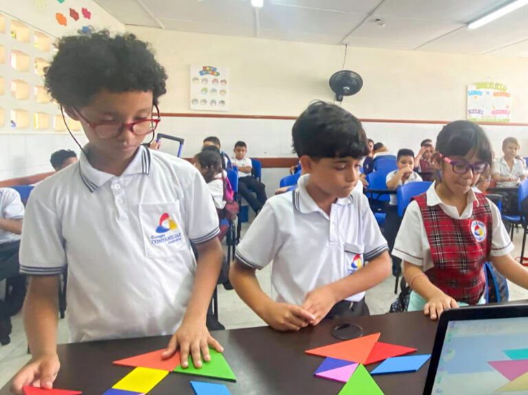 Tres niños en un aula trabajan con figuras de papel de colores. Al fondo, otros estudiantes observan. Predominan tonos azules y blancos en el entorno.