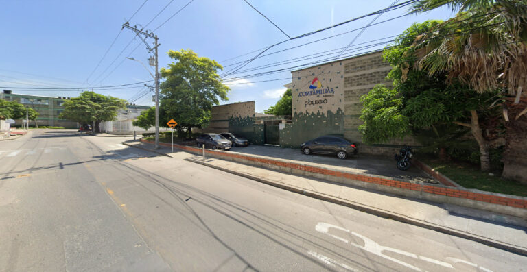 Edificio con logo en la fachada, árboles verdes, vehículos estacionados y una calle despejada bajo un cielo soleado.