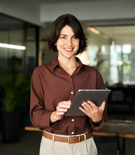 Mujer sonriente con camisa marrón sosteniendo una tablet en un entorno de oficina moderna, con plantas y muebles de trabajo visibles en el fondo.