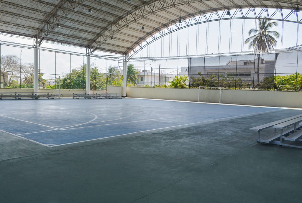 Cancha techada con piso azul y líneas blancas, rodeada de gradas metálicas. Ventanas amplias permiten la entrada de luz natural y se observan árboles en el exterior.