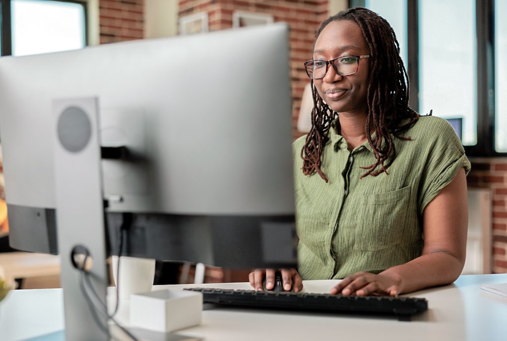Mujer con cabello rizado y gafas, sentada frente a un monitor, usando un teclado en un entorno de oficina con paredes de ladrillo y luz natural. Lleva una camisa verde.
