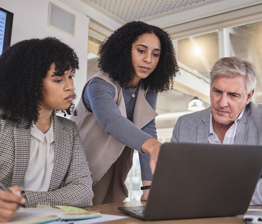 Tres personas en una reunión analizan información en una computadora portátil. Dos mujeres, una con cabello rizado y otra con cabello lacio, y un hombre de cabello canoso. Entorno de oficina moderno.