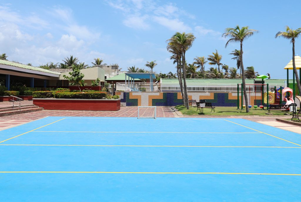 Superficie de juego azul con líneas amarillas, rodeada de palmeras y áreas verdes, edificios en el fondo y un parque infantil visible a la derecha. Cielo despejado.