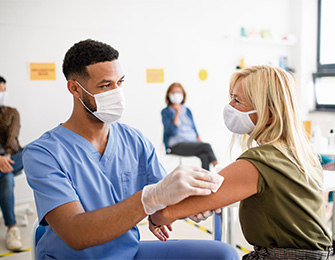 Profesional de la salud administra una vacuna en el brazo de una mujer, ambos usando mascarillas. Al fondo, otras personas esperan en un ambiente clínico. Predominan colores claros.