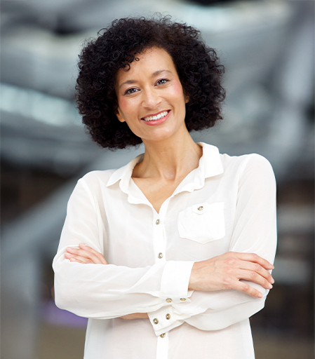 Mujer con cabello rizado y suelto, sonriente, viste una camisa blanca. Se encuentra en un entorno moderno y desenfocado, con colores neutros predominantes.