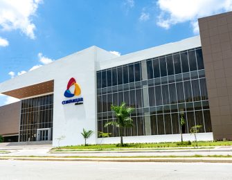 Edificio moderno con fachada de vidrio, logo colorido y áreas verdes alrededor, bajo un cielo parcialmente nublado.