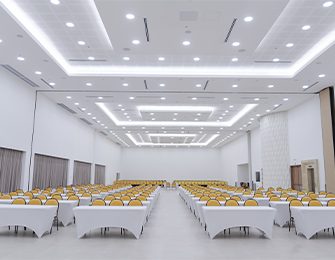 Salón amplio con mesas blancas y sillas amarillas dispuestas en filas, iluminación LED en el techo, paredes claras y cortinas grises, creando un ambiente formal y moderno.