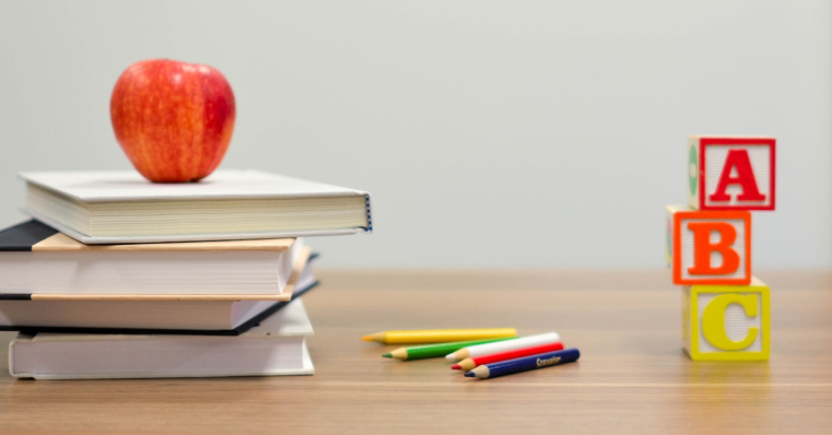 Una manzana roja sobre una pila de libros, junto a bloques de letras coloreados y lápices en diferentes colores sobre una mesa de madera clara.