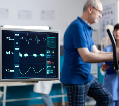 Monitor médico muestra datos de signos vitales, mientras un paciente en ropa cómoda utiliza una máquina de ejercicio, acompañado por dos profesionales de la salud en un entorno clínico.