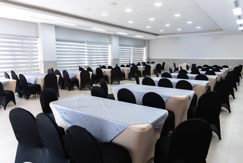 Salón amplio con mesas rectangulares cubiertas de manteles claros y sillas negras. Ventanas con cortinas enrollables permiten la entrada de luz natural. Suelo de cerámica clara.