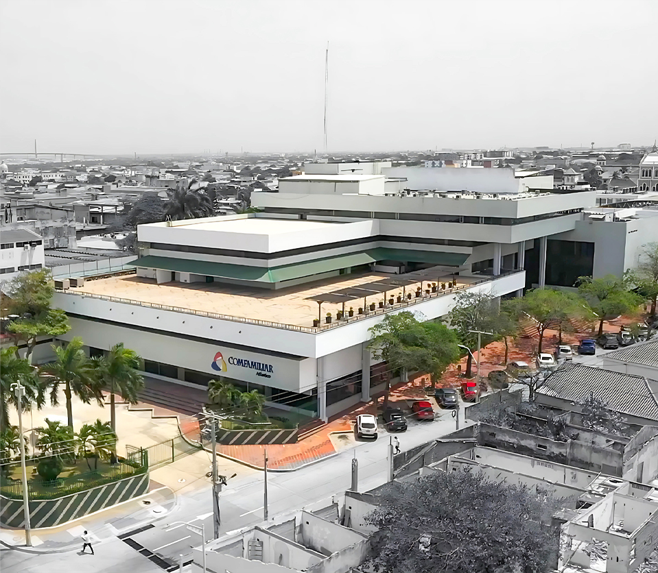 Edificio moderno con varias plantas, rodeado de árboles y vehículos. El logotipo de Comfamiliar es visible en la fachada. Fondo urbano con casas y calles cercanas.