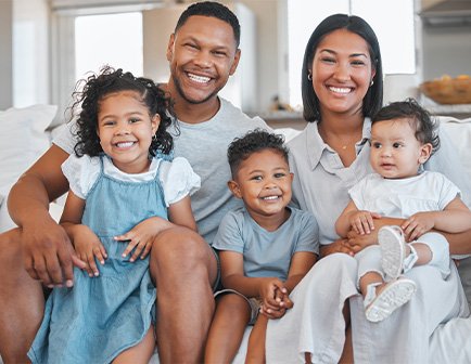 Familia sonriente sentada en un sofá, compuesta por dos adultos y tres niños. Los adultos visten ropa casual clara y los niños llevan diferentes atuendos, todos con expresiones alegres.