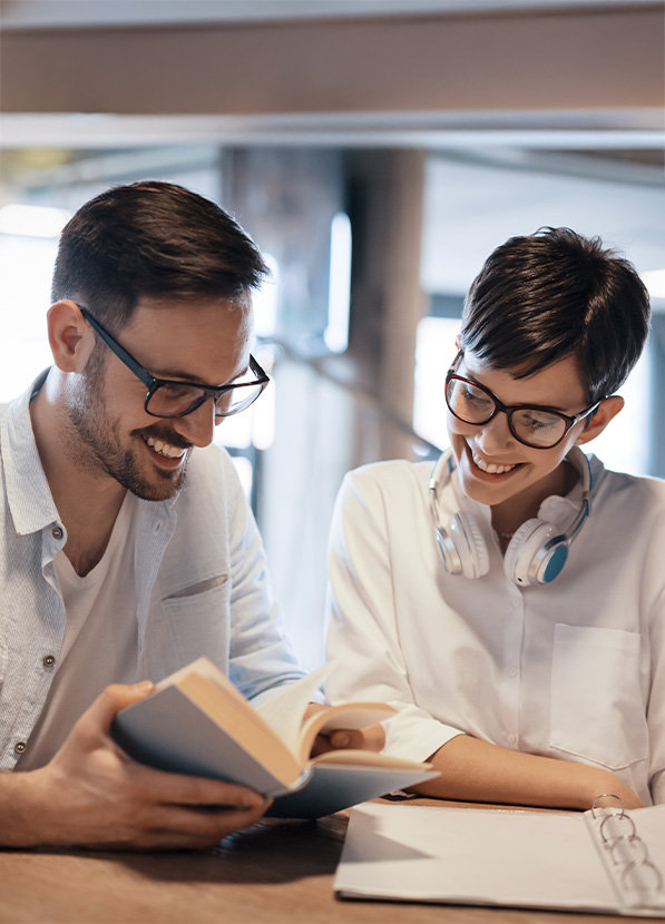 Pareja sonriente revisando un libro en un ambiente de trabajo. El hombre lleva una camisa ligera y gafas, mientras que la mujer usa auriculares y una blusa blanca.