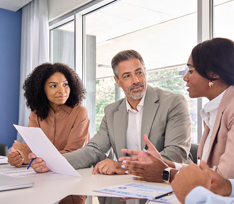 Cuatro personas en una reunión, discutiendo documentos sobre una mesa en un entorno luminoso y moderno.