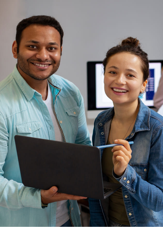 Dos personas sonríen mientras sostienen una computadora portátil y un bolígrafo, en un entorno de oficina con pantallas de computadora en el fondo. Predominan colores neutros.
