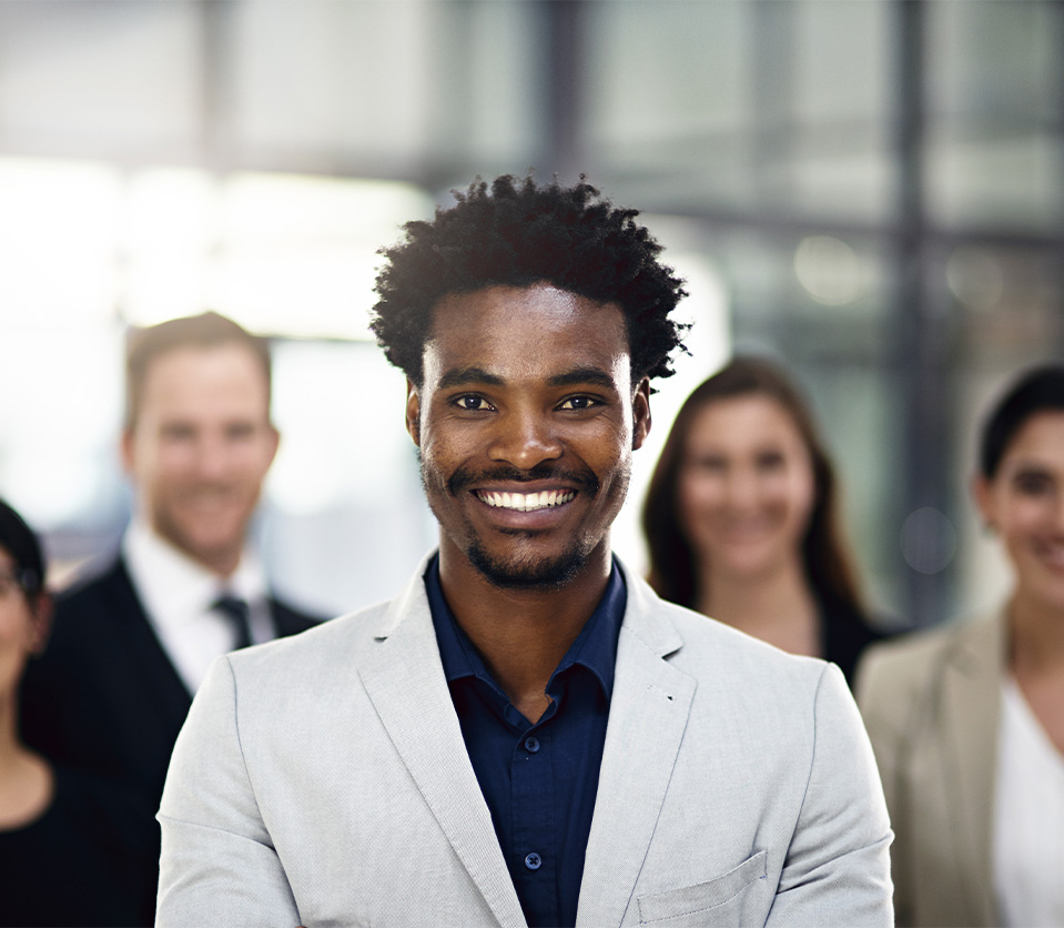 Hombre sonriente en primer plano, con traje claro, rodeado de tres personas en un entorno de oficina moderna.