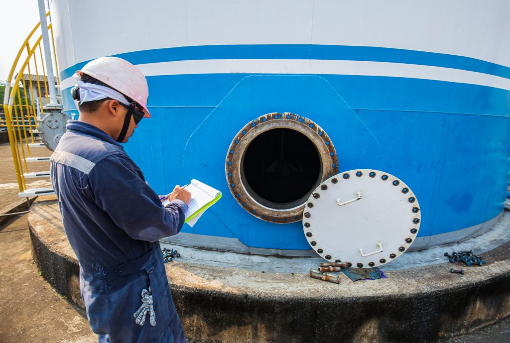 Trabajador con casco y gafas de seguridad, anotando en una libreta frente a un tanque grande azul con una abertura circular y tapa blanca en el suelo. Herramientas y tornillos alrededor.