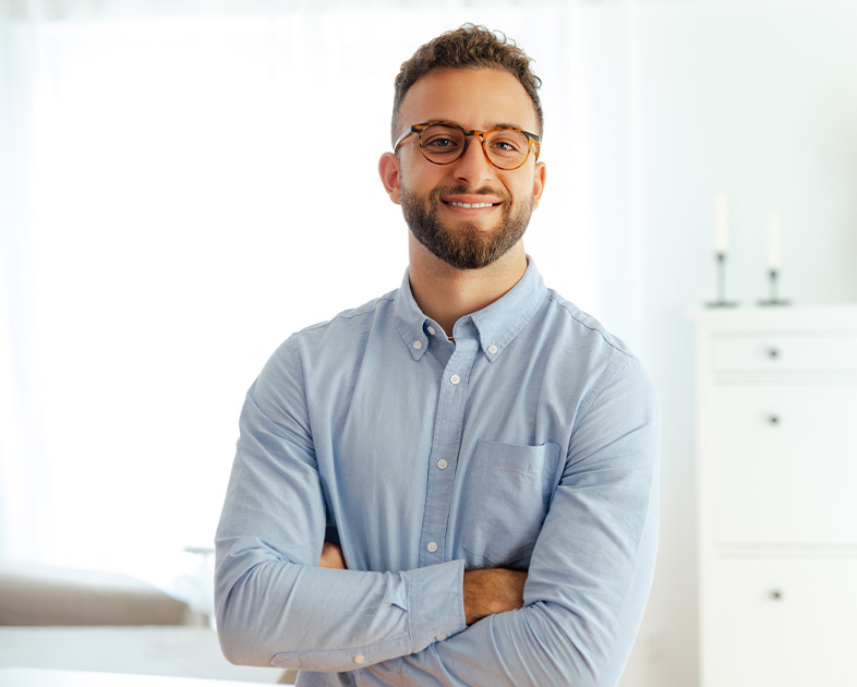 Hombre con gafas y camisa de botones, sonriente, de brazos cruzados, en un entorno luminoso y moderno.