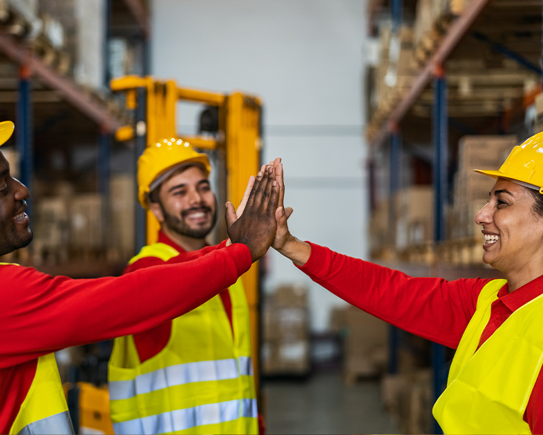 Tres trabajadores en un almacén, vestidos con chalecos de seguridad y cascos, realizan un gesto de celebración, mientras se encuentran entre estanterías llenas de cajas.