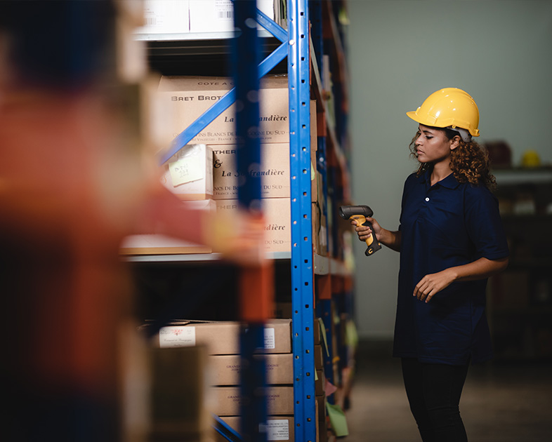 Mujer con casco amarillo utiliza un escáner en un almacén, junto a estanterías llenas de cajas. El entorno es industrial, con colores neutros y iluminación suave.