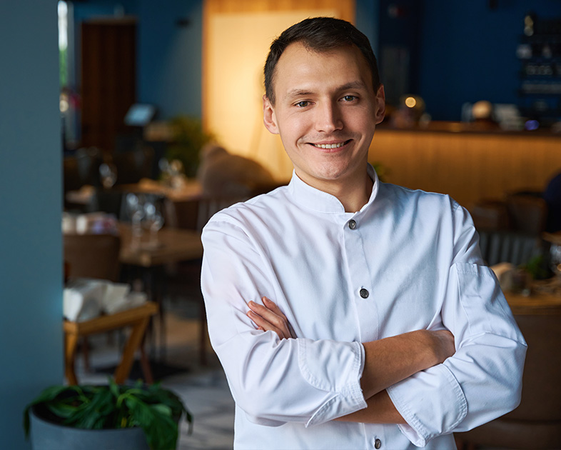 Hombre con camisa blanca sonriente, brazos cruzados, en un entorno de restaurante moderno con decoración acogedora.