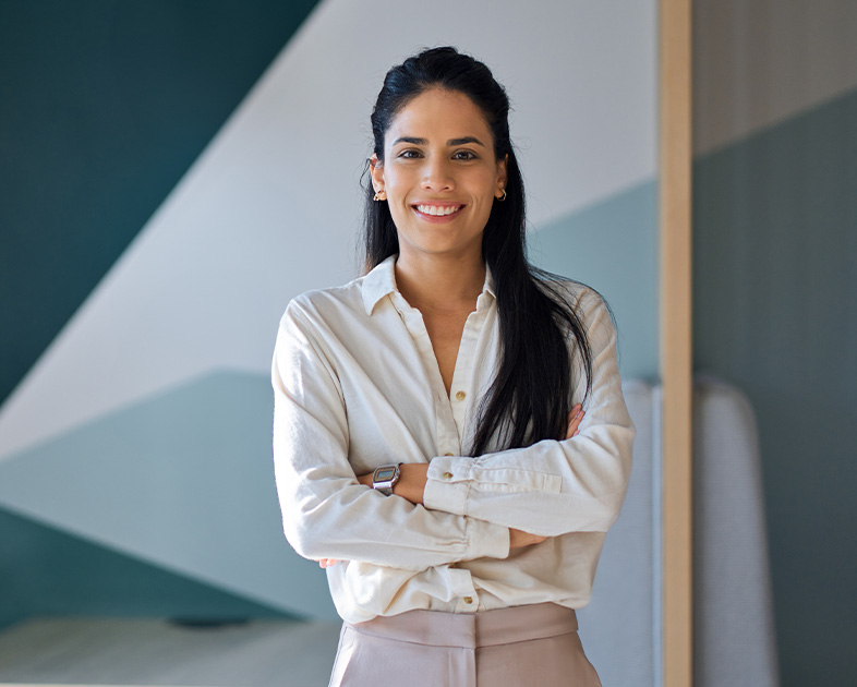 Mujer sonriente con camisa blanca y pantalones claros, de pie en un entorno moderno con colores suaves.