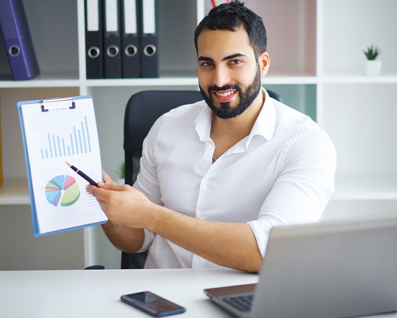 Hombre con barba sonriente sostiene un informe con gráficos en una oficina moderna, con computadora portátil y teléfono móvil en la mesa. Fondo con estanterías y planta decorativa.
