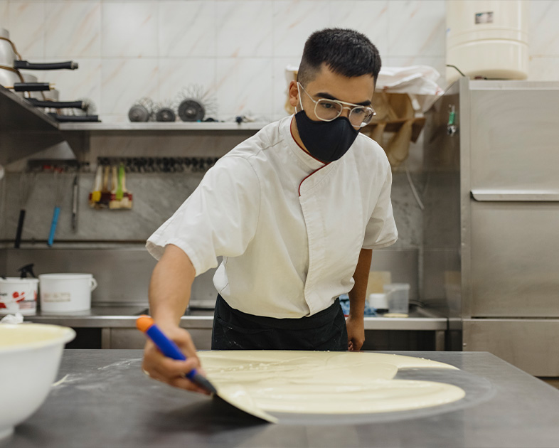 Chef con mascarilla trabaja la masa en una cocina, utilizando una espátula sobre una superficie metálica.