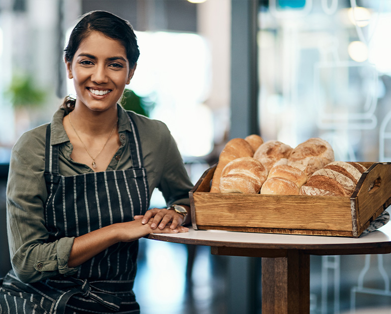 Mujer sonriente con delantal de rayas, apoyada en una mesa, junto a una bandeja de pan recién horneado en un entorno luminoso y moderno.