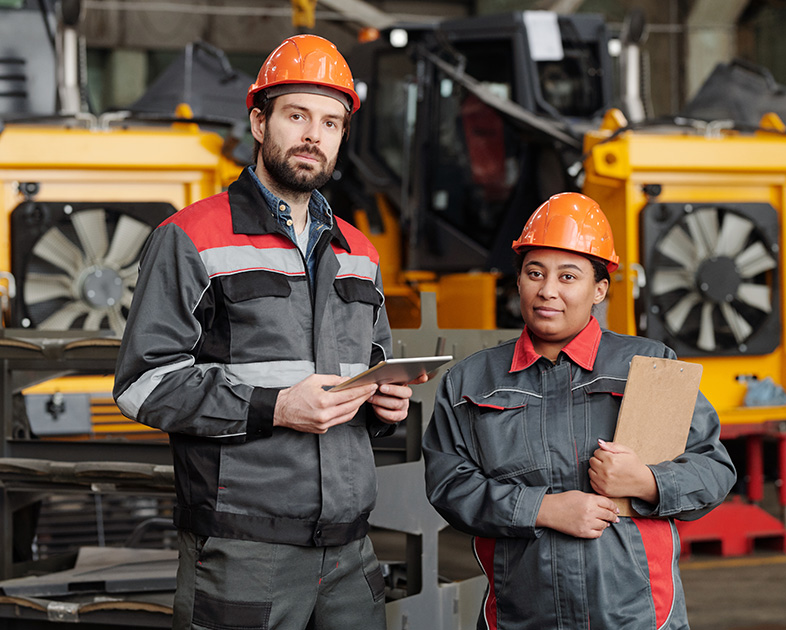 Dos trabajadores en un entorno industrial, vistiendo overoles grises y cascos naranjas, sosteniendo tabletas y carpetas, con maquinaria pesada y herramientas de fondo.