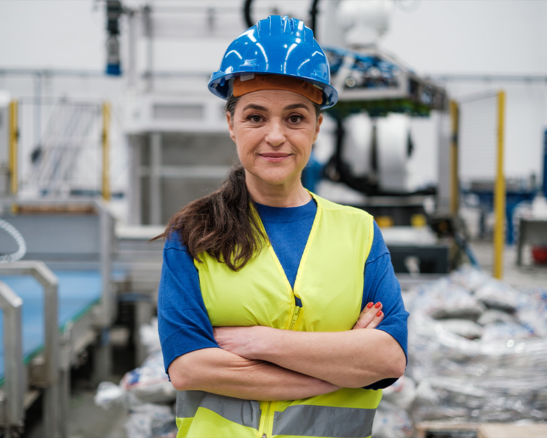 Mujer con casco azul y chaleco amarillo en un entorno industrial, con brazos cruzados y una expresión seria, rodeada de maquinaria y materiales en un fondo de fábrica.